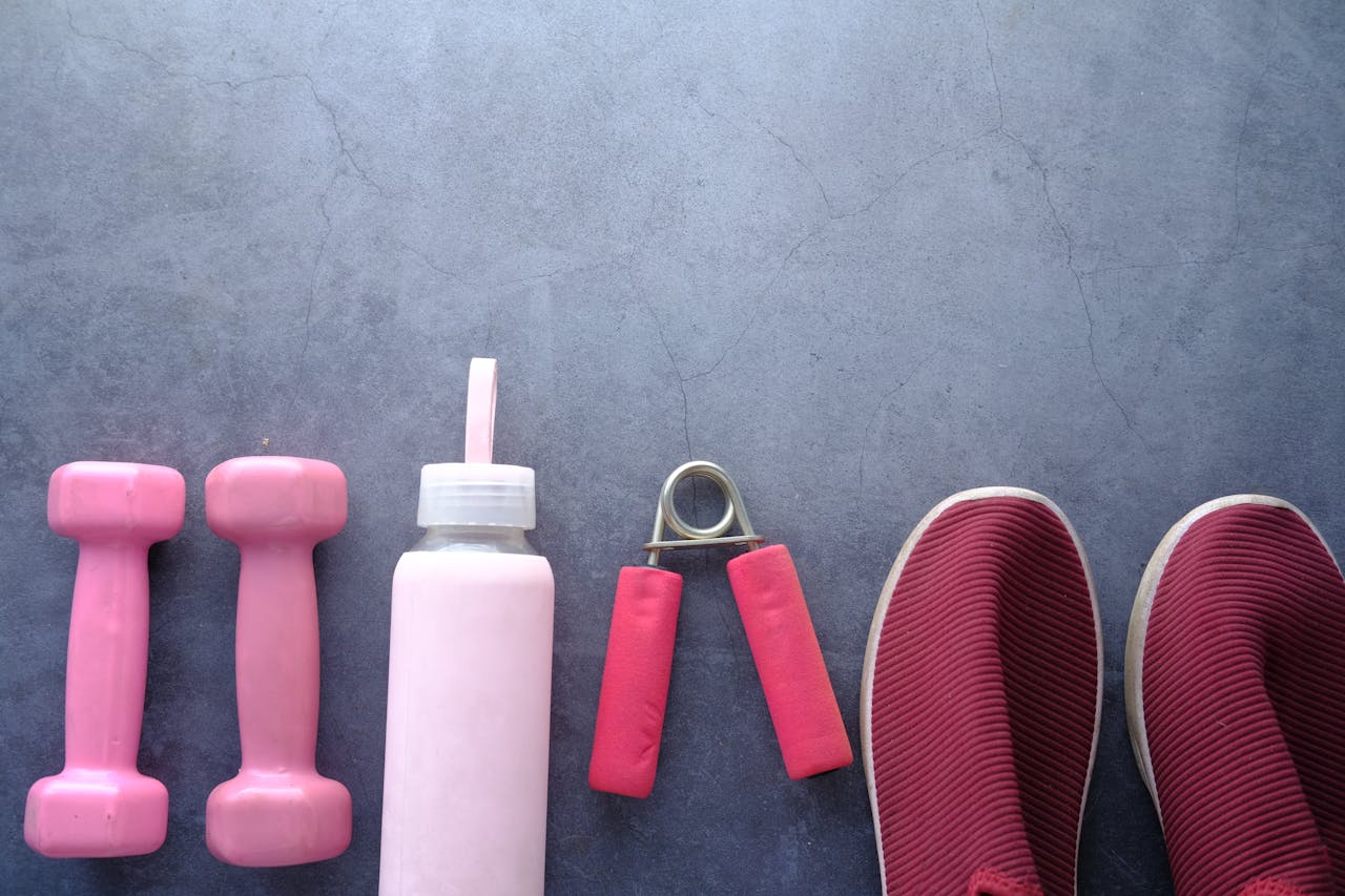 Flat lay of pink fitness equipment and shoes on a gray surface including dumbbells, water bottle, and grip strengthener.